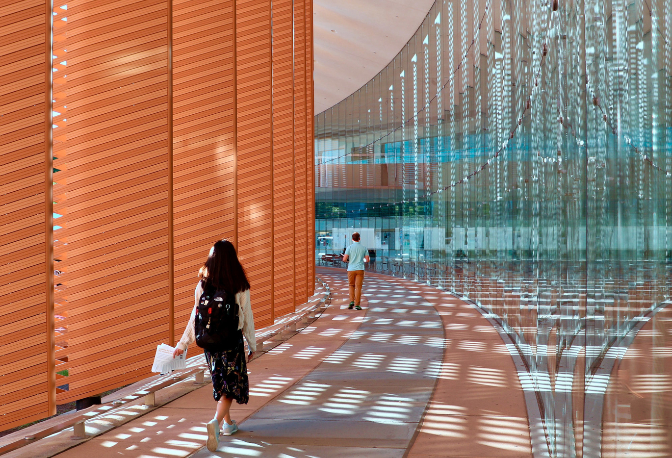 "Two people are walking away on a pathway, with tall glass windows on the right, and shaders on the left."