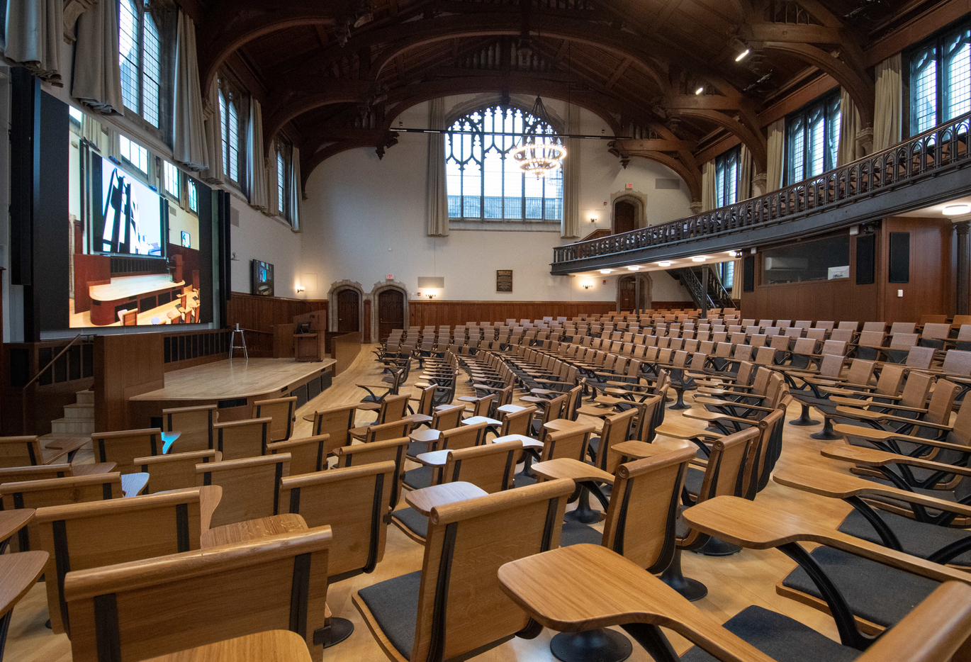 "Interior of a lecture hall. Wooden desks with chairs are looking at a large projector screen. There is a second floor of desks above and to the right. Beautiful wooden arches are attached to the ceiling"