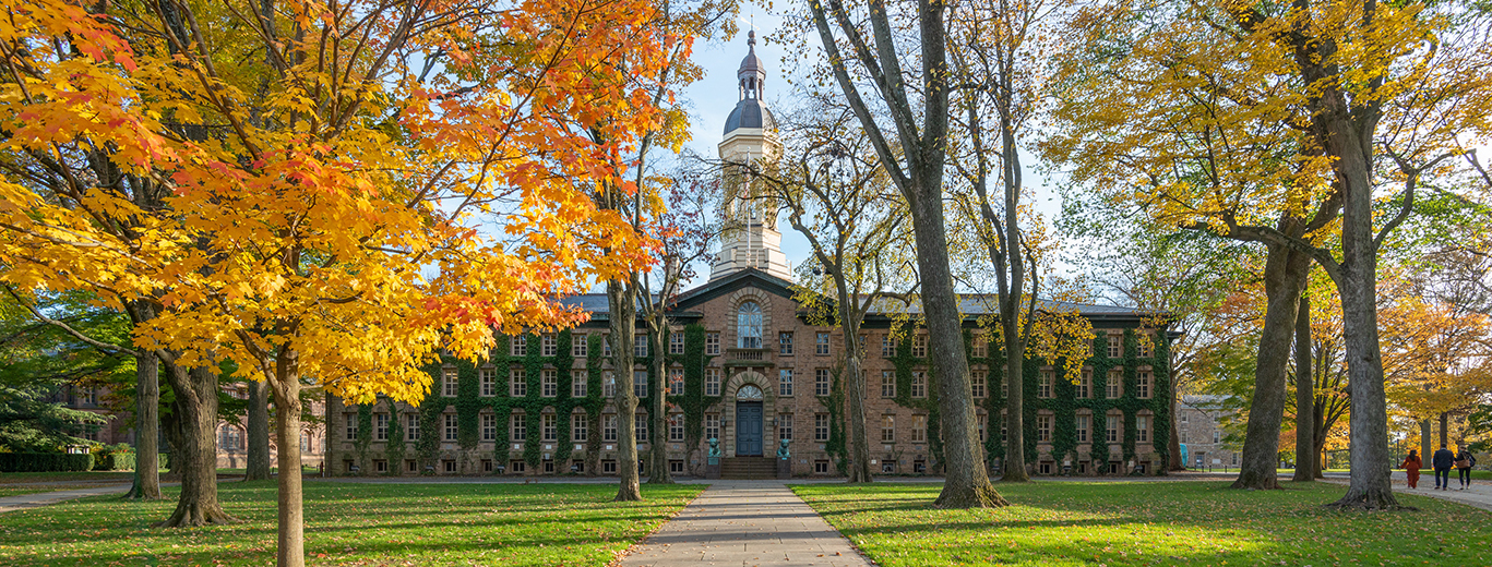 "Exterior of Nassau Hall. A paved path is in the centre towards the entrance, with grass and trees on either size. The building is brick with three floors, and a white spring rising in the centre"