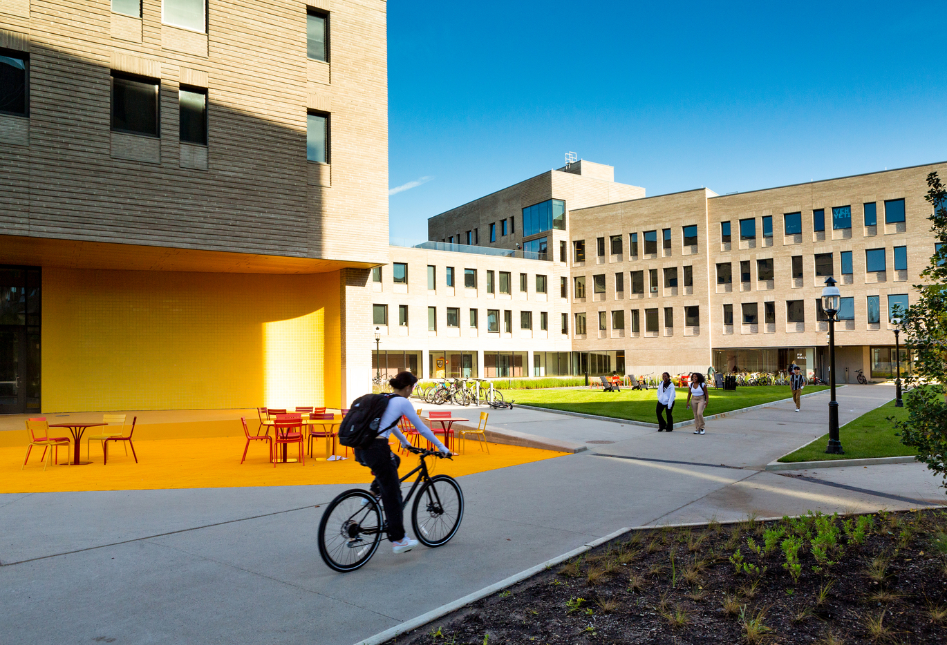 "External view, grey buildings are in the rear, with a grassy area and paths in front. People are sitting on the grass, walking and cycling on the paths. A painted orange seated area with tables is to the left."